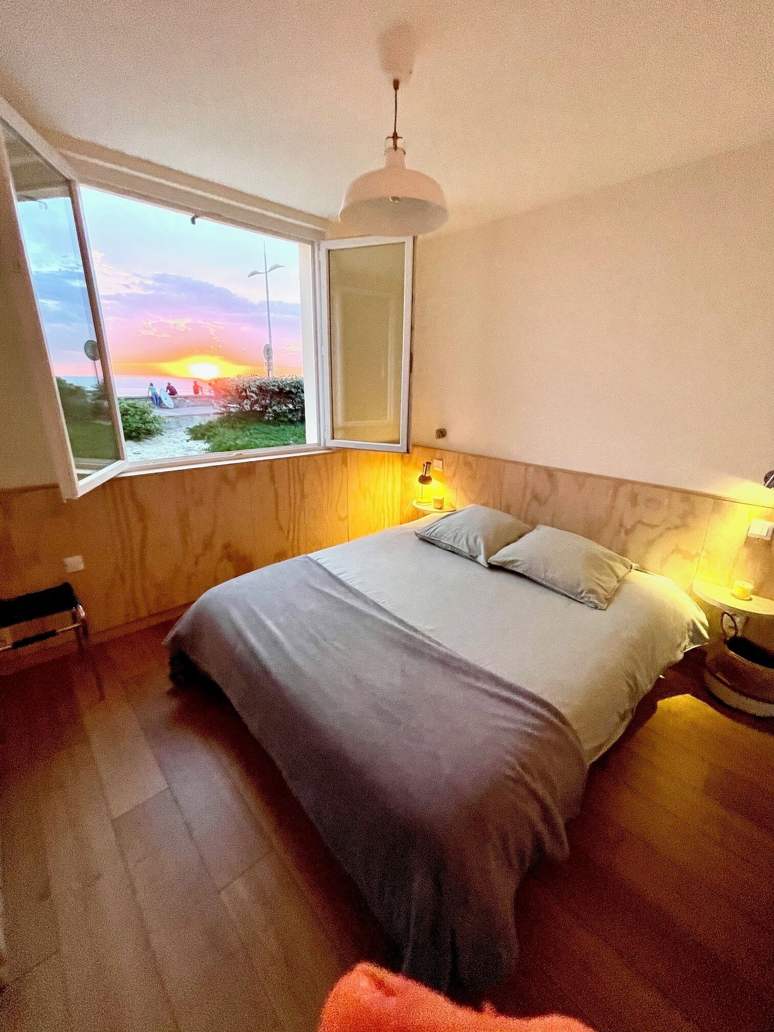 Une chambre avec une grande fenêtre, un lit, une table de chevet avec une lampe, et une vue sur le coucher de soleil.