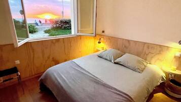 Une chambre avec une grande fenêtre, un lit, une table de chevet avec une lampe, et une vue sur le coucher de soleil.