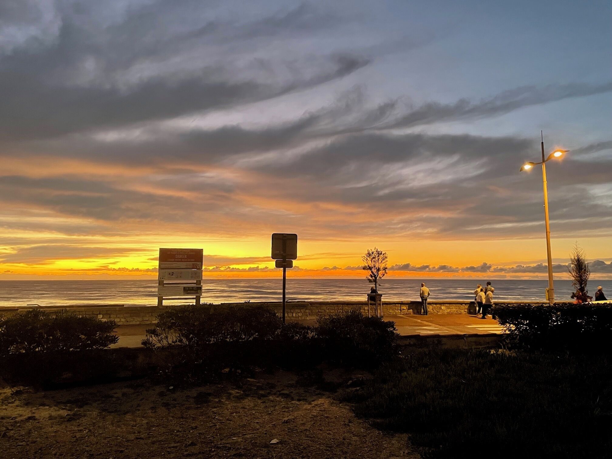 A sunset view with a beach, a warning sign, and a few people walking.
