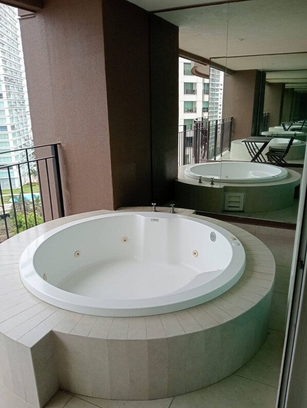 A circular hot tub on a balcony with a view of a building and a pool in the background.