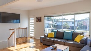 Uma sala de estar com sofá, piso de madeira, TV fixada na parede e uma grande janela com vista para casas.