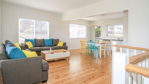 Ampla sala de estar com sofá cinzento, mesa de centro de madeira e mesa de jantar com cadeiras coloridas.