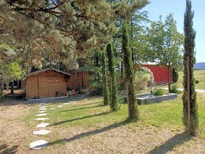 Une cabane en bois rustique, accessible par un sentier de pierres, entourée de grands arbres et avec un bâtiment rouge en arrière-plan.