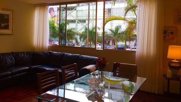 A hotel room with a dark leather sofa, a glass dining table with chairs, a chandelier, and a large window with a view of palm trees and buildings. - "Your home in Miraflores" 4 pers (Lima)