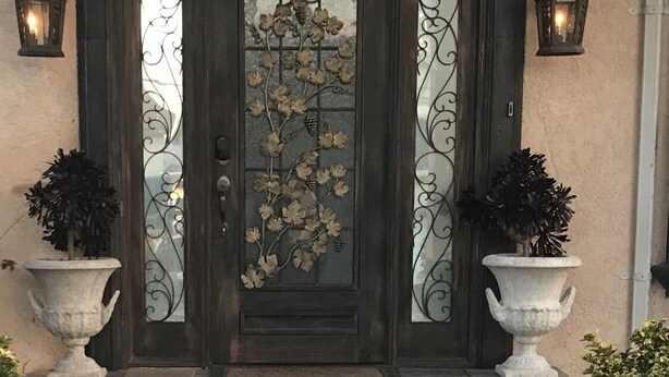 Eine dunkle Holztür mit dekorativem Glasfenster, beidesseits je ein Blumentopf und je eine Wandleuchte.