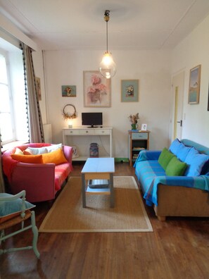 A living room with a wooden floor, a white table, two sofas with colorful cushions, a TV on a white stand, and a hanging light fixture.
