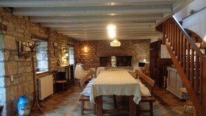 Une pièce rustique avec un mur en pierre, une cheminée, une table en bois et des bancs, ainsi qu’un escalier.