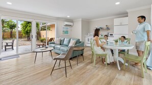 Una sala de estar moderna con una mesa de comedor de vidrio, un sofá y una zona de cocina con armarios blancos y un refrigerador.