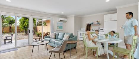 A modern living room with a glass dining table, a sofa, and a kitchen area with white cabinets and a refrigerator.