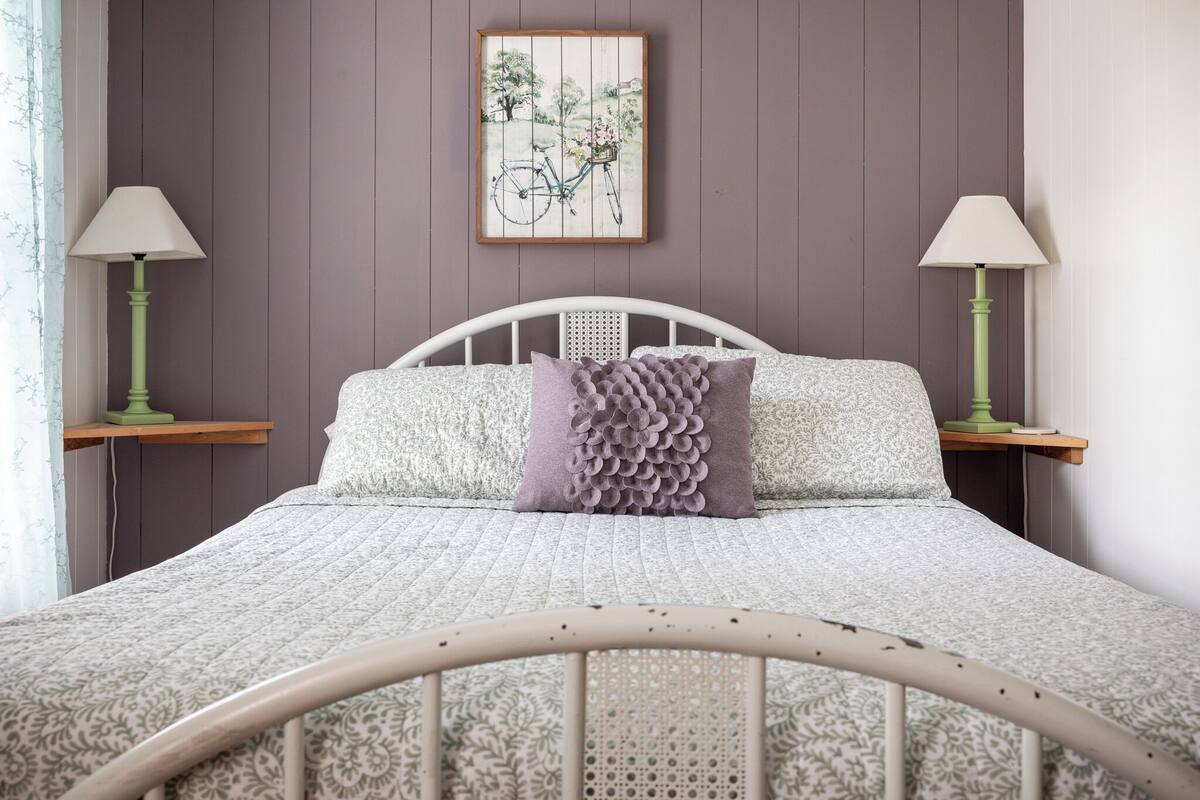 A bedroom with a white metal bed frame, a patterned bedspread, two nightstands with green lamps, and a framed picture of a bicycle on the wall.