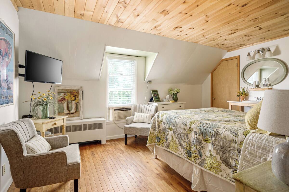 A bedroom with a wooden ceiling, a bed with a floral bedspread, a television, a window with blinds, and a small table with a vase of flowers.