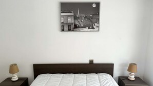 Ein Schlafzimmer mit einem Bett, zwei Nachttischen, einer Lampe auf jedem Tisch, einer weißen, gesteppten Bettdecke und einem Schwarz-Weiß-Stadtbild an der Wand.