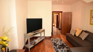 Ein modernes Wohnzimmer mit einem Flachbildfernseher auf einem Lowboard, einer Couch mit Kissen, Holzboden und einem Flur, der zu einer Tür führt.