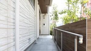 A narrow corridor with a wooden fence on one side and a white brick wall on the other. There are metal handrails along the corridor, and a small window is visible in the distance. - GRAND BASE Hakata City (Fukuoka)