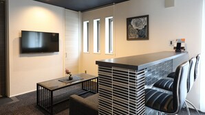 A modern living room with a flat-screen TV, a coffee machine, and a bar area with cushioned chairs. - GRAND BASE Hakata City (Fukuoka)