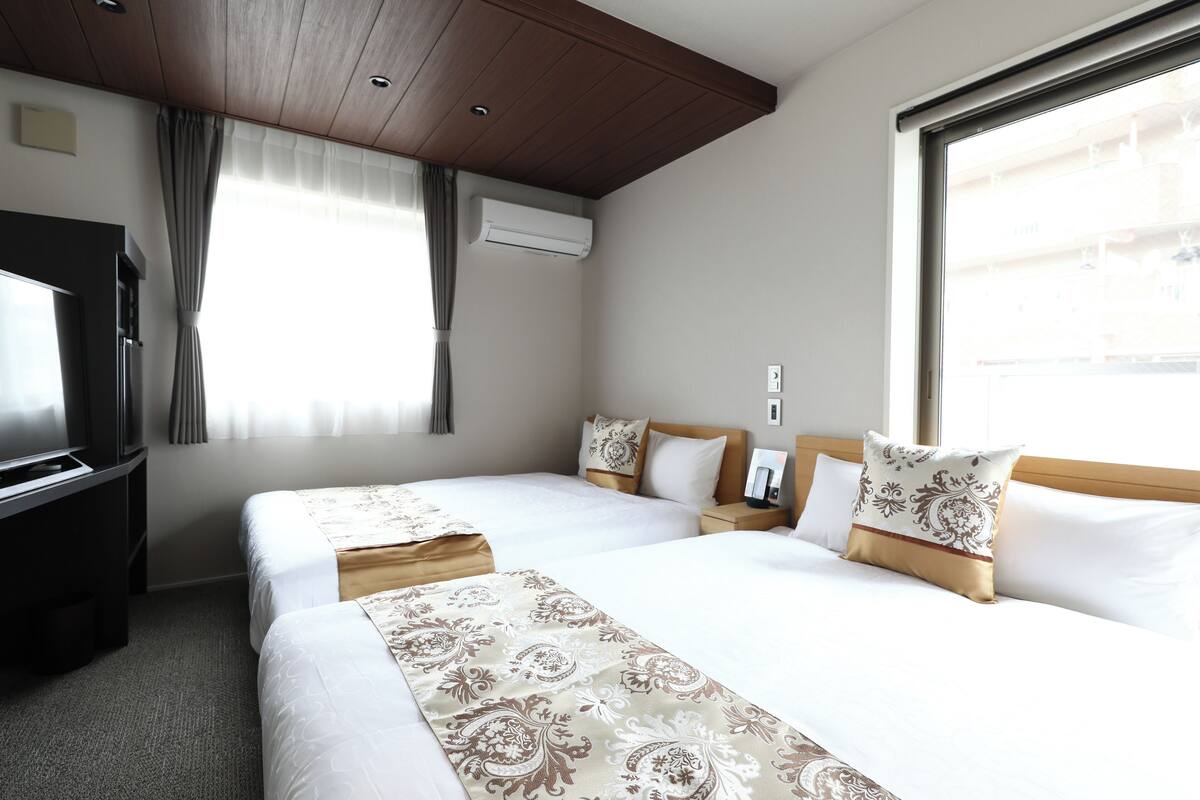 A hotel room with two beds, a TV, an air conditioner, and a window with curtains.