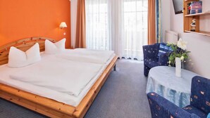 Habitación de hotel con cama doble, una mesita con un florero, una silla y un televisor sobre una estantería.