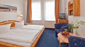 Habitación de hotel con cama de madera, una mesita con fruta, un sofá, un televisor y una estantería con libros.