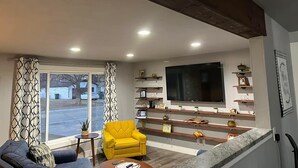 Una sala de estar con un sofá, un sillón amarillo, un televisor y estantes con objetos decorativos.