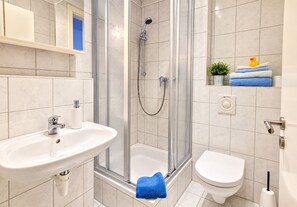 A bathroom with a shower, sink, toilet, and a small shelf with towels and a rubber duck. - Room 5 - Pension and Apartments Haus Antje (Heringsdorf)