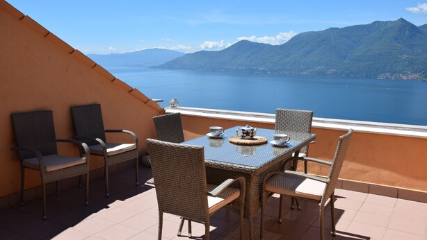 Une terrasse dotée de meubles en osier et d’une table dressée pour deux personnes, offrant une vue sur un lac paisible et des montagnes.