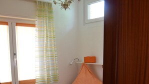 Une petite chambre avec un lit simple, une tête de lit en bois, une couvre-lit à motifs, un rideau rayé vert et blanc, un oreiller orange, une petite fenêtre et un mur blanc agrémenté d’un luminaire.