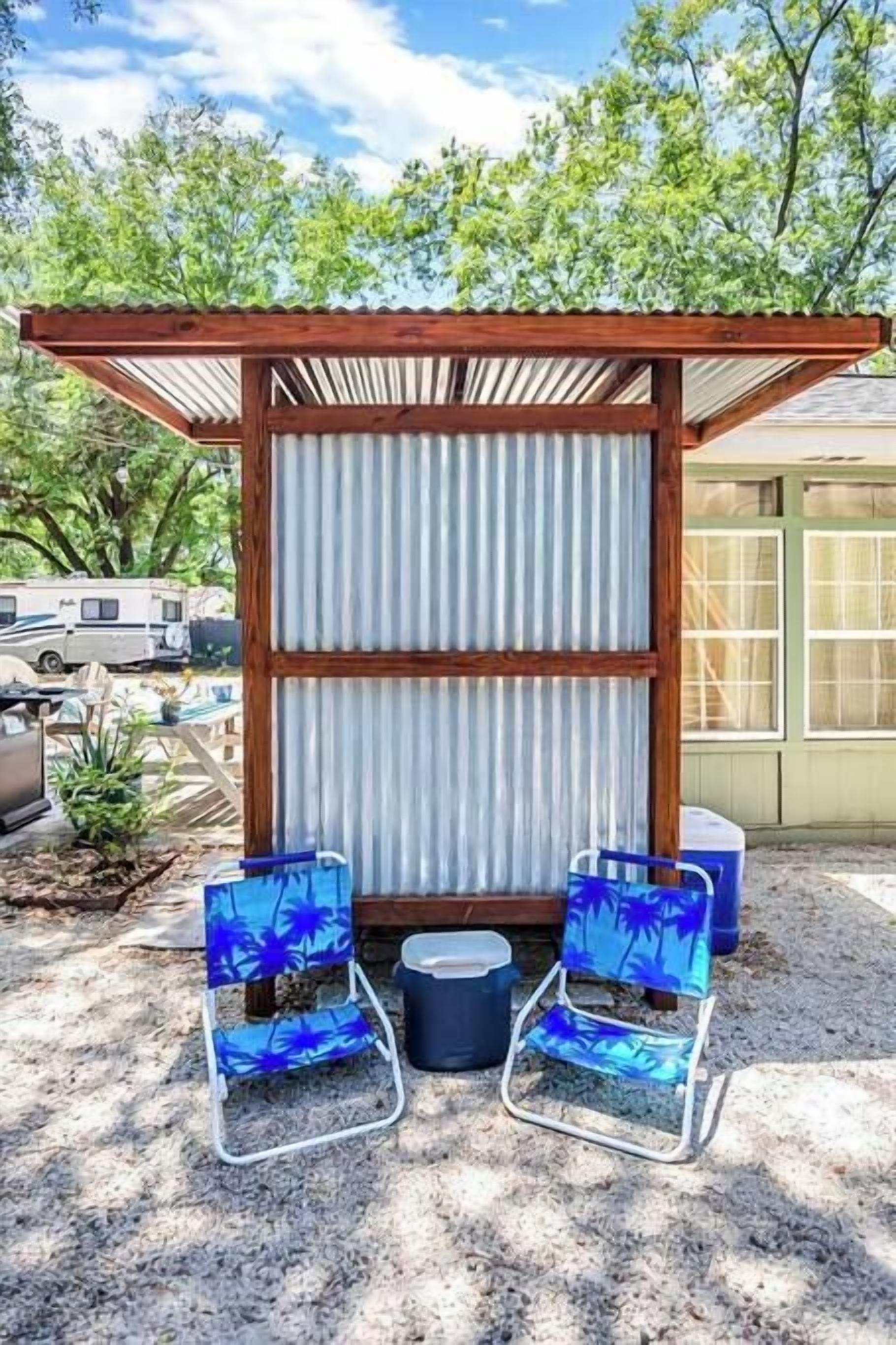 A covered outdoor seating area with two chairs and a table, surrounded by a corrugated metal fence and wooden beams.
