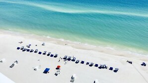 Une vue aérienne d’une plage avec des rangées de transats et de parasols, un océan bleu limpide et une plage de sable.