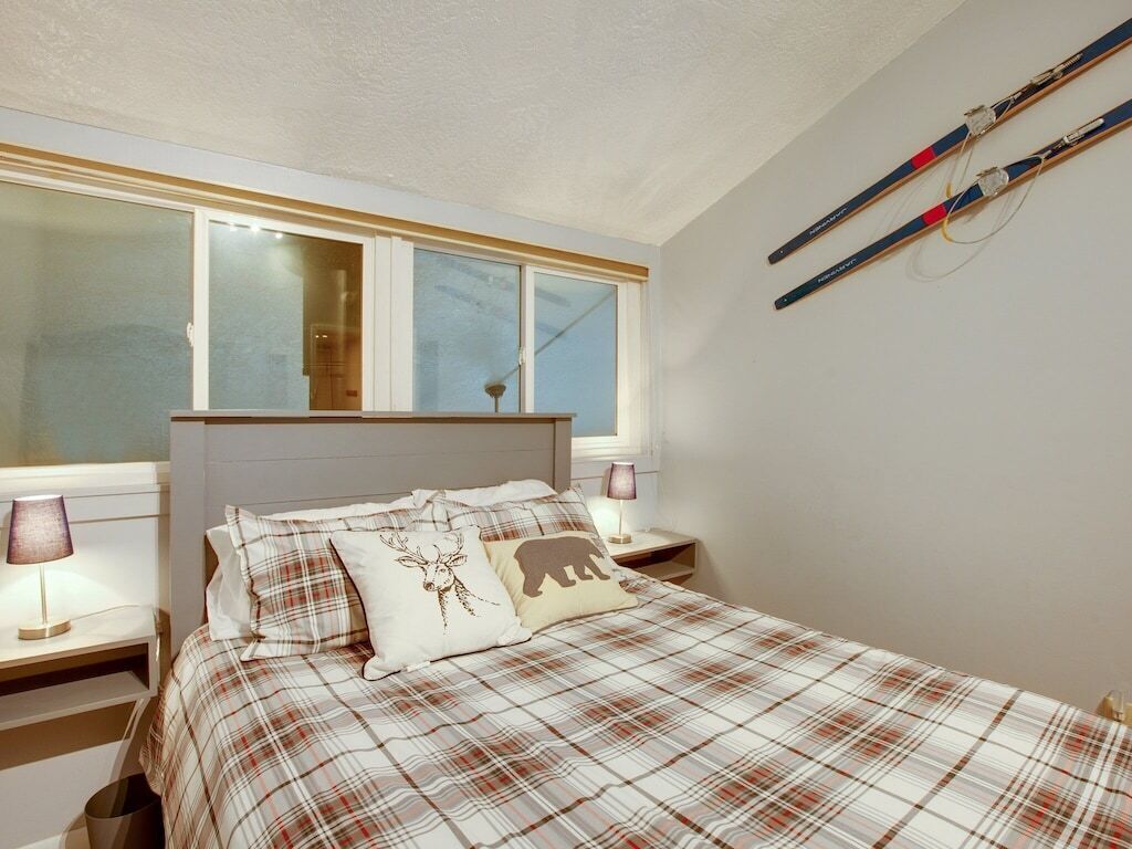 A bedroom with a plaid bedspread, a headboard, two bedside lamps, a window with blinds, and a wall-mounted ski rack.