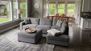 A spacious living room with a gray sectional sofa, a dining area with wooden chairs, and a kitchen with white cabinets. - Woodinville Wine Country New Custom Home - Seasonal Pricing - Casa Bambino (Woodinville)
