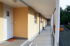 A covered corridor with a tiled floor, white door, and windows with flower boxes.