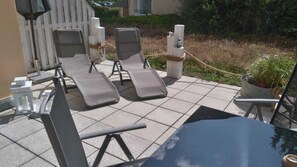 Un espace patio comprenant deux chaises longues et une table, entouré d’une clôture blanche et de plantes en pot.
