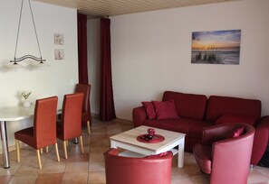 Uma sala de estar com um sofá vermelho, poltronas a combinar, uma mesa de centro branca e um quadro de um pôr do sol.