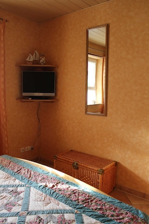 A bedroom with a bed, a television on a shelf, a mirror, and a wicker chest of drawers.