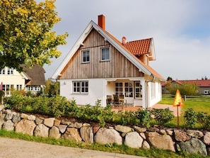 A wooden house with a sloped roof, a stone wall in front, and a grassy yard.