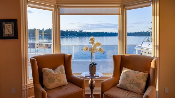 A room with two leather armchairs facing a large window overlooking a lake, a small table with a vase of flowers, and a grill outside the window. - Stellamar on Hayden, Close-in Executive Waterfront (Hayden)