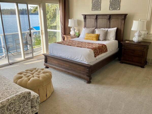A bedroom with a large bed, two bedside lamps, a nightstand, a tufted ottoman, and a view of a lake through a sliding glass door.