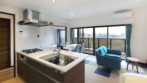 A modern kitchen with a built-in stove, sink, and ventilation hood. There is a dining area with a table and chairs, and a balcony with a view of the city. - Grand Residence Hakataeki Minami (Fukuoka)