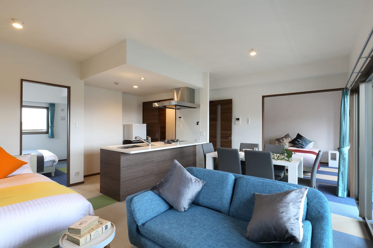 A modern hotel room with a bed, a blue sofa, a dining area, and a kitchenette.