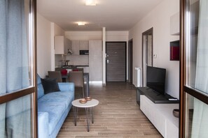 Un espace de vie moderne comprenant un canapé, une petite table ronde, un téléviseur et, au fond, un coin cuisine.