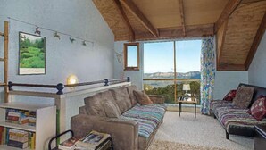 A cozy living room with a sofa, a small table, and a bookshelf. There is a large window offering a view of the outdoors. - Stunning Cliff Face and Valley Views (KATOOMBA)