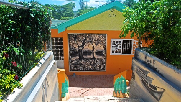 Un carreró estret amb un edifici petit que presenta un mural de paisatge amb arbres i una barca, envoltat de vegetació i un paviment de rajoles.