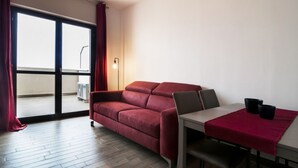 Een kamer met een rode bank, een eettafel met twee stoelen, een rode plaid op de tafel en een groot raam met gordijnen.