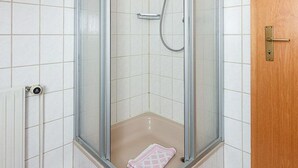 Een douche met glazen wanden, een handdouche, een handdoek op de vloer en een houten deur aan de rechterkant.