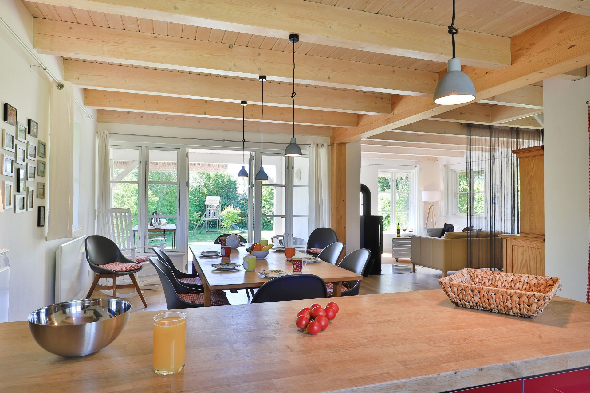 A spacious kitchen with wooden beams, a dining area with chairs, a large island with a bowl of tomatoes, and a view of the outdoors.