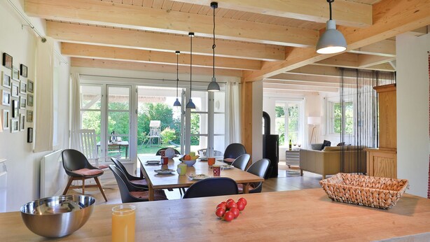 Une cuisine spacieuse avec des poutres en bois, un coin repas avec des chaises, un grand îlot central sur lequel se trouve un saladier rempli de tomates, et une vue sur l’extérieur.