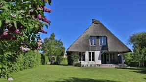 Une maison à toit de chaume, aux murs blancs, entourée d’une pelouse bien entretenue et de plantes en fleurs.
