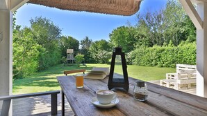 Un espace de restauration en plein air avec une table en bois, un toit de chaume et une vue sur un jardin agrémenté d’une structure de jeu en bois et d’une lanterne.