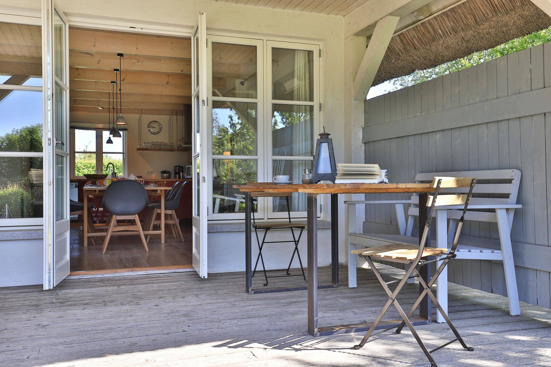 Une terrasse ouverte avec une table et des chaises en bois, une lanterne, et une vue sur la cuisine et la salle à manger.