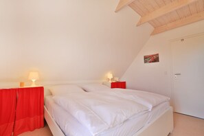 Um quarto com teto inclinado, duas mesas de cabeceira com luminárias, uma cama com roupa de cama branca, uma cortina vermelha e uma porta com um pequeno quadro.
